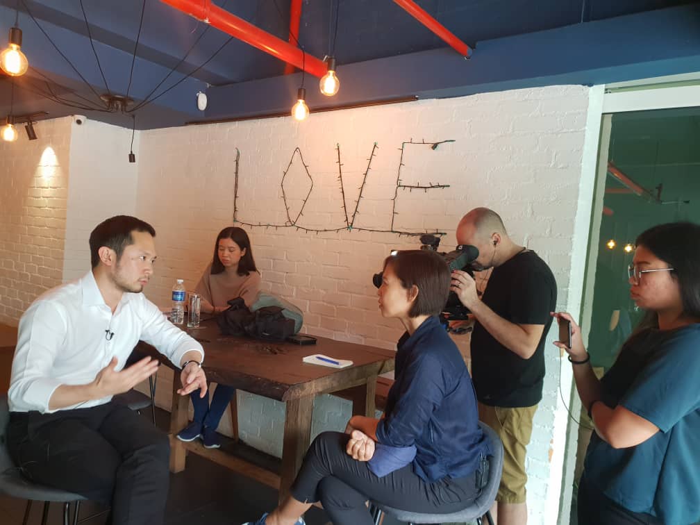 Chan Eu Boon being interviewed on camera with a white LOVE backdrop and film crew.